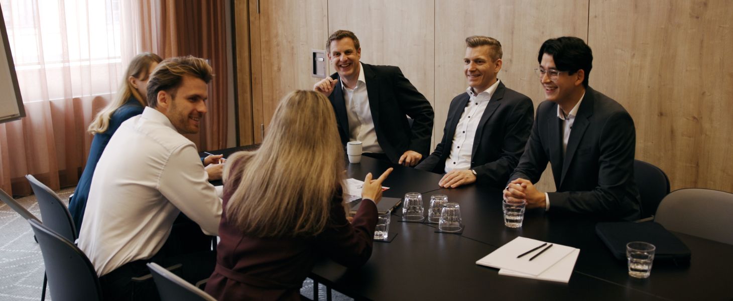 Gruppe von Business-Professionals sitzt gemeinsam an einem Konferenztisch und diskutiert in entspannter, positiver Atmosphäre, symbolisch für Teamarbeit und Karriere bei Consileon