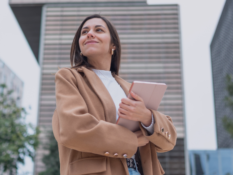 Selbstbewusste Frau im Business-Outfit steht vor moderner Architektur und hält ein Tablet in den Armen, symbolisch für moderne Karrierewege