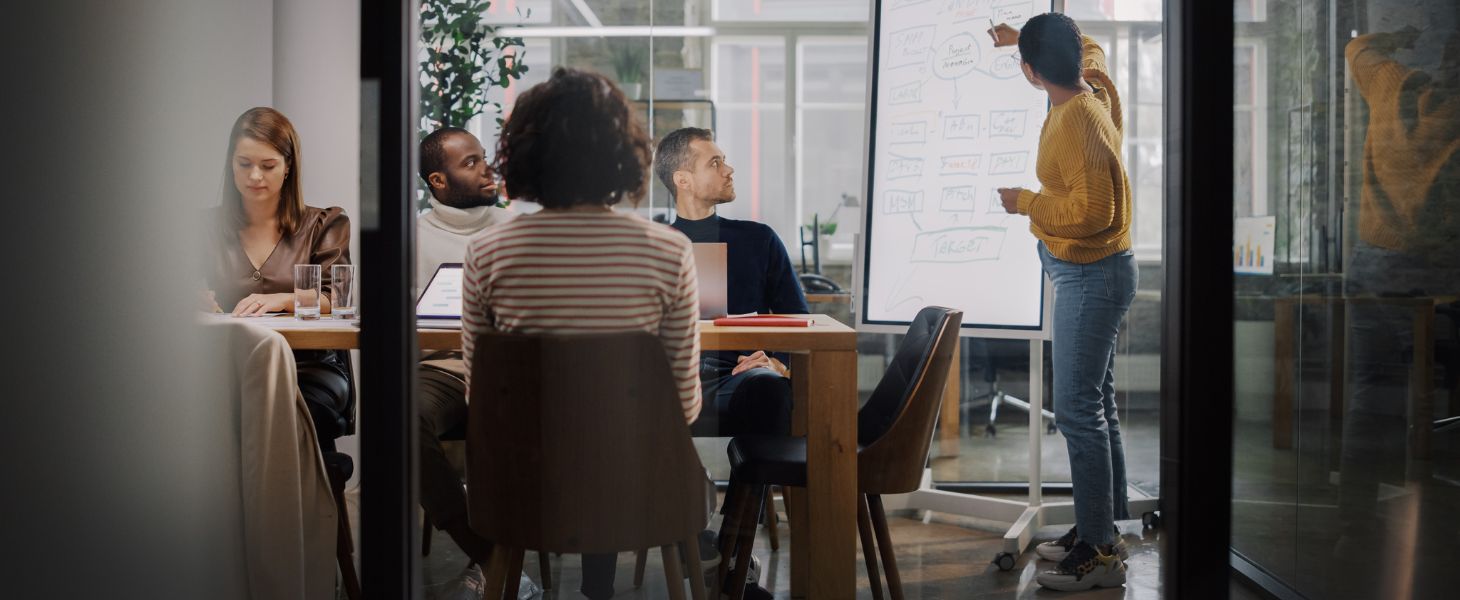 Teammeeting in einem modernen Büro: Eine Person präsentiert am Whiteboard, während Kolleginnen und Kollegen am Tisch zuhören, symbolisch für strategische Planung zum Marketing-Automation