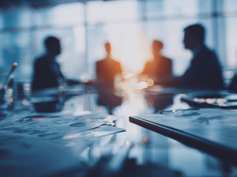 Unscharfe Silhouetten von Personen in einem Meeting mit Unterlagen im Vordergrund, symbolisch für strategische Abstimmung und Zusammenarbeit im Corporate Banking.