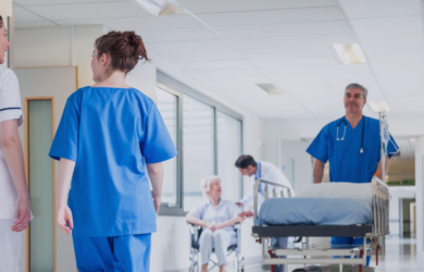 Pflegepersonal und Ärzte im Krankenhausflur, Patient im Rollstuhl und Bett im Hintergrund, symbolisch für Zusammenarbeit und Versorgung im Gesundheitswesen.