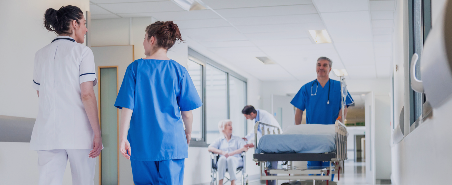 Pflegepersonal und Ärzte im Krankenhausflur, Patient im Rollstuhl und Bett im Hintergrund, symbolisch für Zusammenarbeit und Versorgung im Gesundheitswesen.